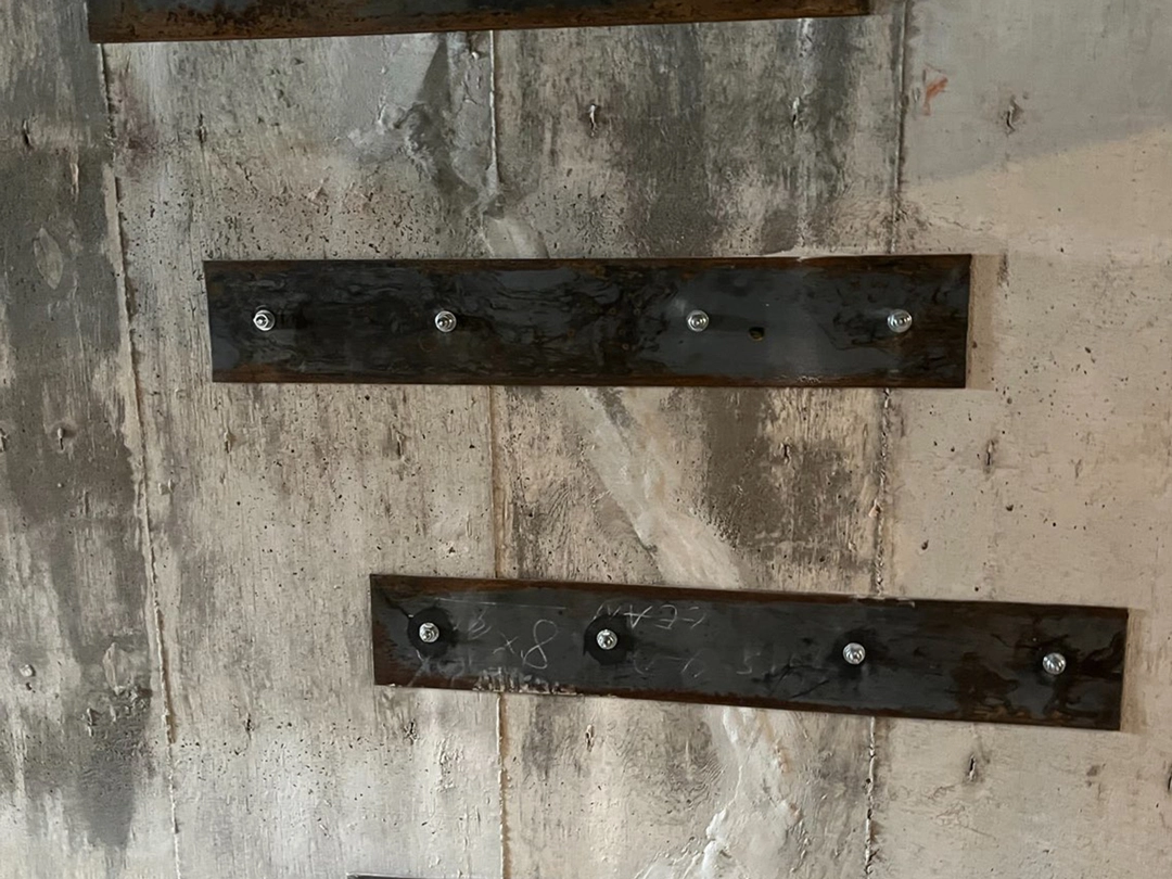Rusty metal plates affixed to a textured concrete wall, each plate secured with visible screws, showcasing a weathered industrial aesthetic