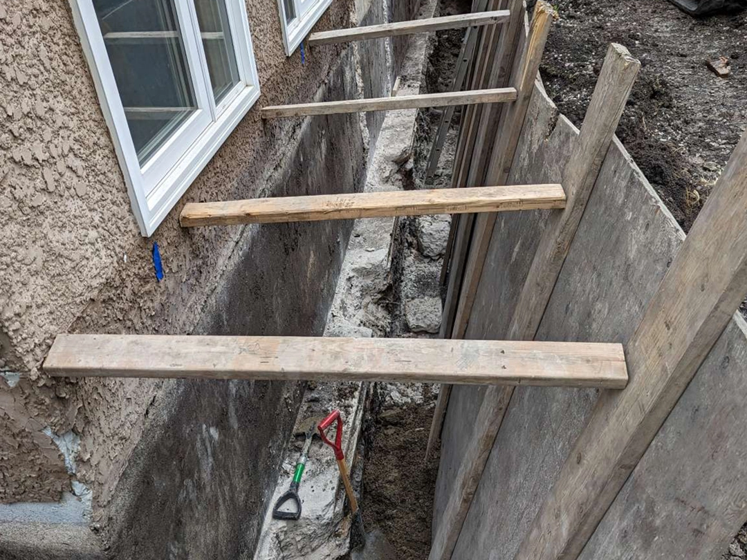 Wooden planks extend over a trench next to a house, with tools visible on the ground and unfinished concrete walls in the background