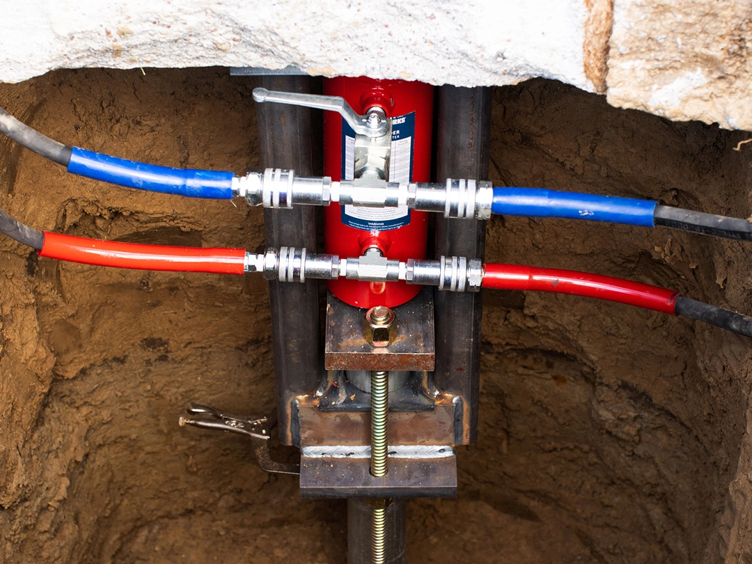 Close-up of a red hydraulic pump in a trench, with blue and red hoses connected to it, set in earthy surroundings