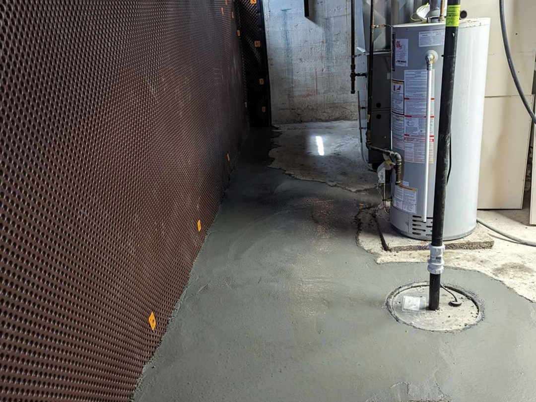 Freshly poured concrete floor in a basement with a water heater and drainage pipe nearby, against a textured brown wall