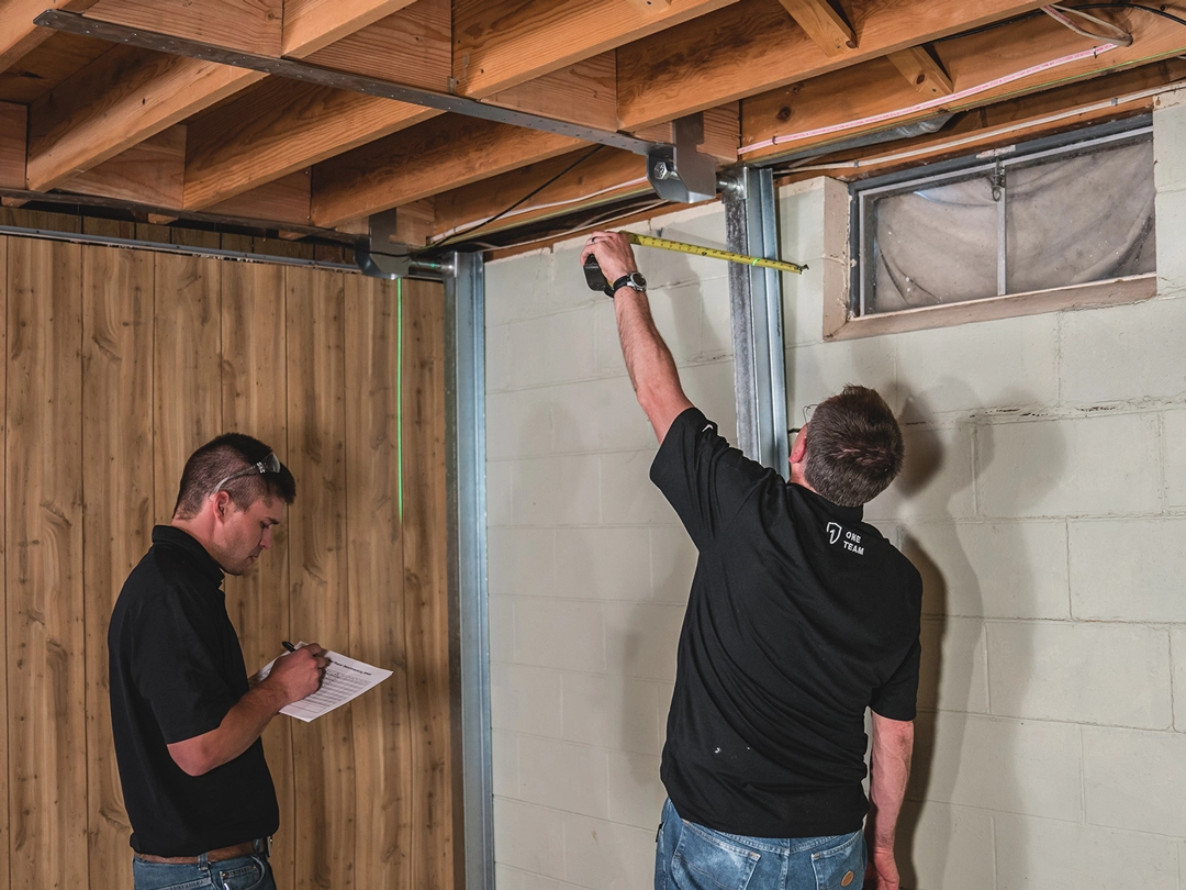 Two men in black shirts are measuring and inspecting a basement area, with one holding a measuring tape and the other taking notes